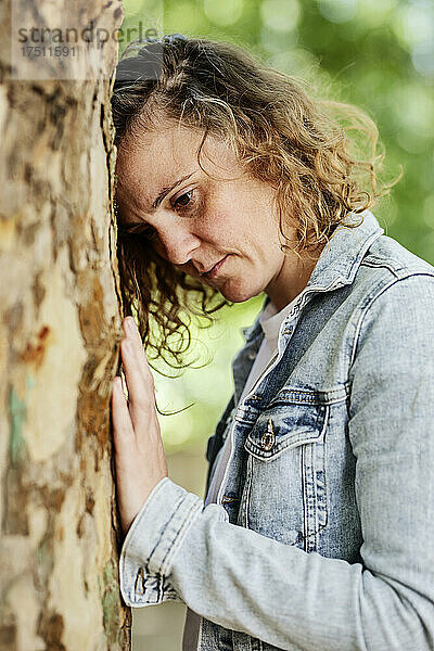 Nahaufnahme einer nachdenklichen Frau in Jeansjacke  die sich im Park an einen Baumstamm lehnt