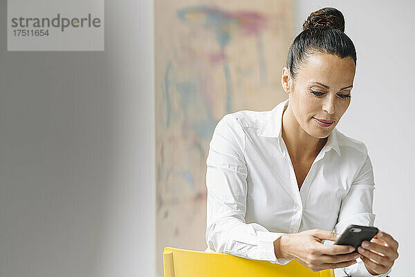 Unternehmerin nutzt Mobiltelefon  während sie im Heimbüro auf einem Stuhl an der Wand sitzt