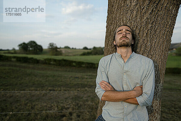 Entspannter junger Mann lehnt an einem Baumstamm auf dem Land