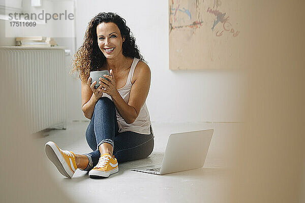 Lächelnde schöne Frau hält eine Kaffeetasse in der Hand  während sie zu Hause einen Laptop auf dem Boden benutzt