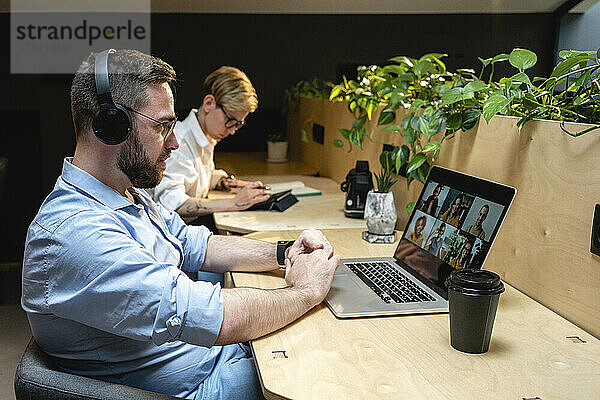Geschäftsmann führt Videoanruf mit Kollegen über Laptop  während er neben einer Geschäftsfrau am Schreibtisch im Kreativbüro sitzt