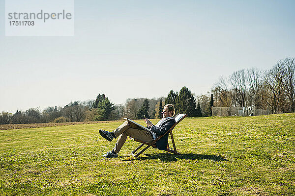 Reifer männlicher Berufstätiger benutzt Smartphone  während er an sonnigen Tagen auf einem Stuhl mit Solarpanel im Park sitzt