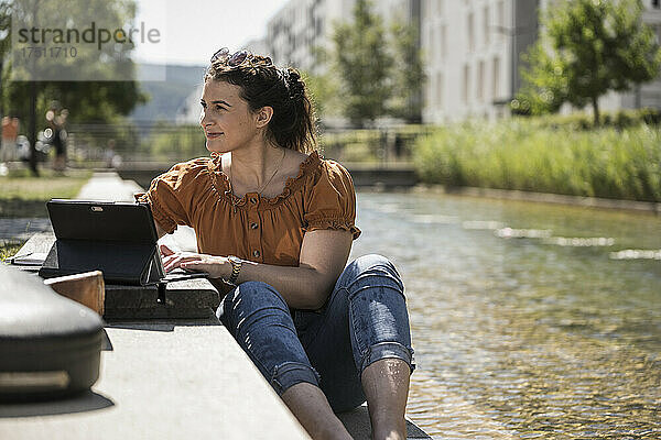 Nachdenkliche Frau mit Laptop sitzt an sonnigen Tagen am Teich im Park