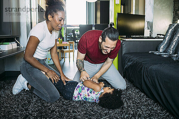 Verspielte Eltern kitzeln ihre Tochter  die zu Hause auf dem Teppich im Wohnzimmer liegt