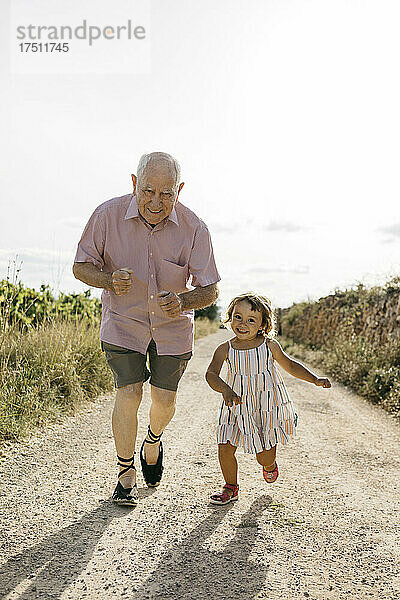 Verspielter älterer Mann rennt an sonnigen Tagen mit seiner Enkelin auf einer unbefestigten Straße inmitten von Pflanzen