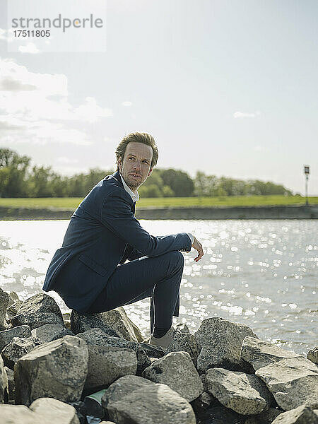 Nachdenklicher Geschäftsmann sitzt an sonnigen Tagen auf einem Felsen am Flussufer vor dem Himmel