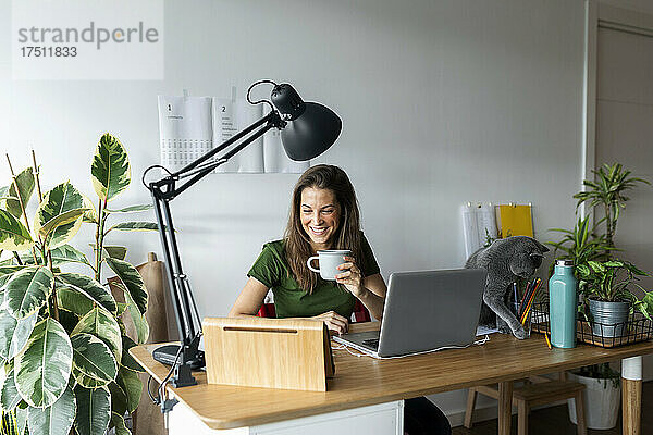 Lächelnde Unternehmerin hält Tasse und digitales Tablet auf dem Schreibtisch im Heimbüro