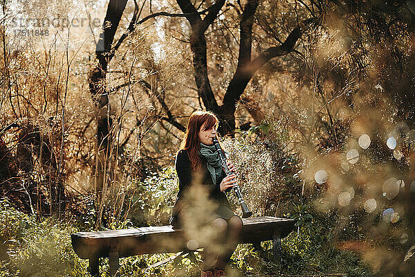 Junge Frau sitzt auf einer Bank und spielt Musikinstrument vor kahlen Bäumen im Wald