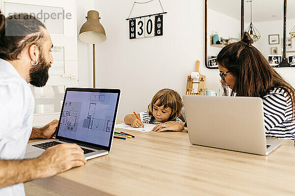 Eltern mit Laptops schauen ihrer Tochter zu  wie sie auf dem Tisch im Esszimmer malt