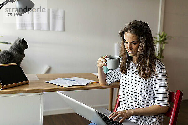 Seriöse Geschäftsfrau hält Tasse und Laptop in der Hand  während sie zu Hause auf einem Stuhl sitzt