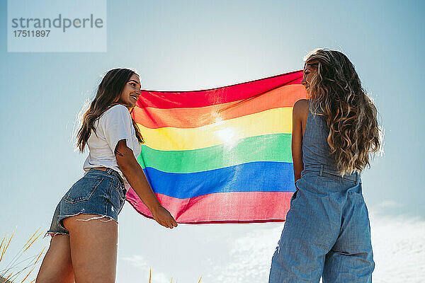 Zwei lächelnde junge Frauen halten eine LGBT-Flagge unter blauem Himmel