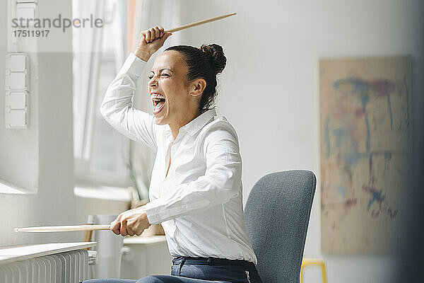 Fröhliche Geschäftsfrau hält schreiende Trommelstöcke in der Hand  während sie im Heimbüro auf einem Stuhl sitzt