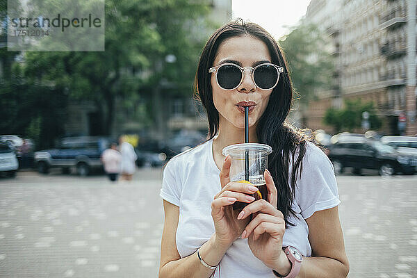 Junge Frau mit Sonnenbrille trinkt Erfrischungsgetränk  während sie in der Stadt steht