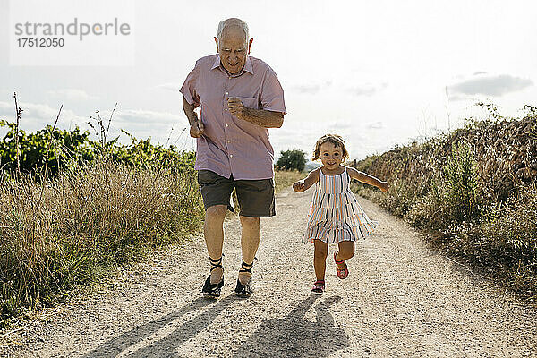 Verspielter älterer Mann rennt mit Enkelin auf unbefestigter Straße inmitten von Pflanzen