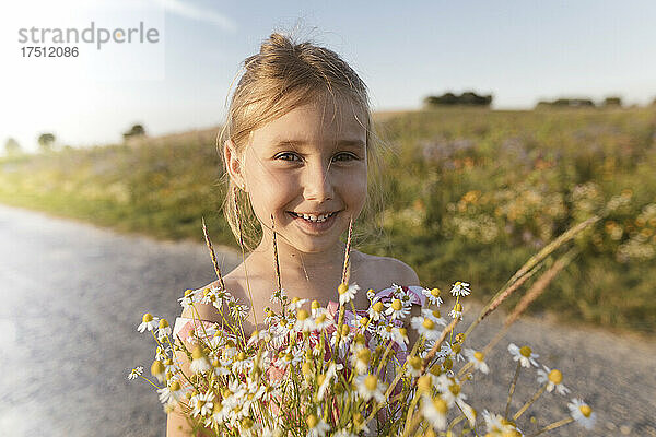 Nahaufnahme eines lächelnden Mädchens  das bei Sonnenuntergang Blumen auf der Straße vor dem Himmel hält