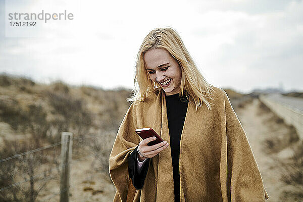 Lachende junge Frau schaut am Strand auf ihr Smartphone
