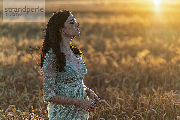 Junge Frau mit geschlossenen Augen steht bei Sonnenuntergang inmitten von Weizenfeldern auf einem Bauernhof