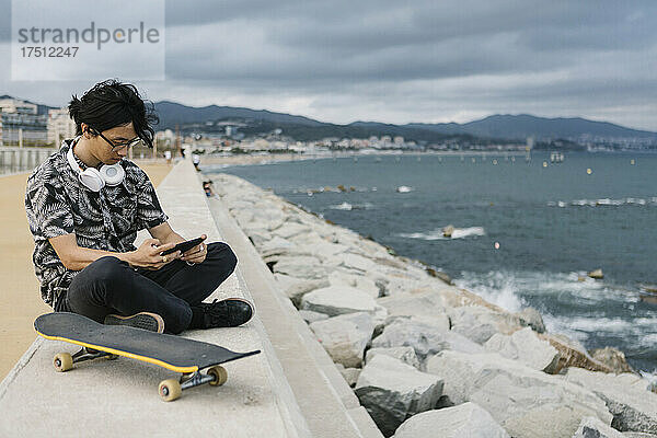 Junger Mann benutzt Smartphone  während er mit Skateboard auf Stützmauer am Meer sitzt