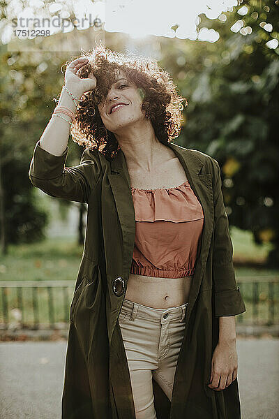 Lächelnde Frau mit lockigem Haar  die einen Mantel trägt  während sie im Park steht