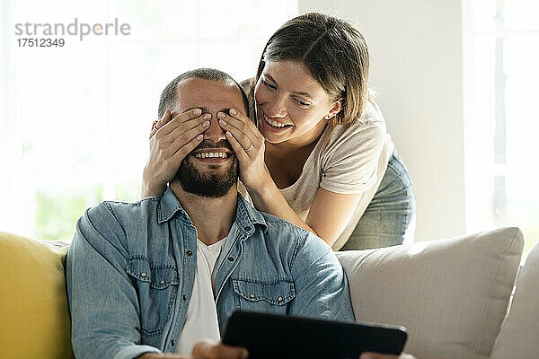 Junge Frau überrascht Mann und bedeckt seine Augen  während er auf der Couch sitzt