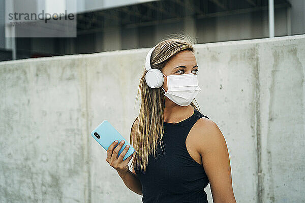 Junge athletische Frau mit Schutzmaske  Kopfhörer und Smartphone