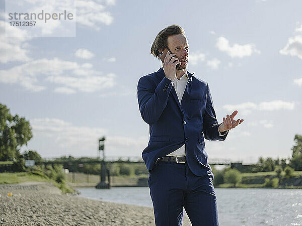 Männlicher Unternehmer spricht über Smartphone  während er am Flussufer vor dem Himmel steht