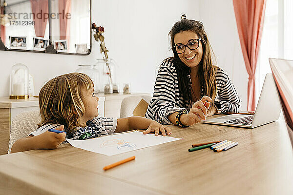 Lächelnde Mutter mit Laptop und Blick auf die Tochter  die auf dem Tisch im Esstisch malt