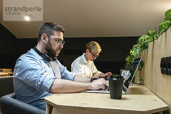 Geschäftsmann benutzt Laptop  während er mit einer Kollegin am Schreibtisch im beleuchteten Coworking-Büro arbeitet