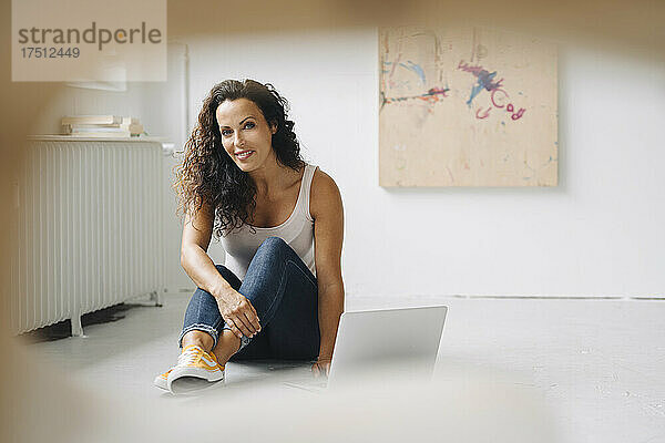 Lächelnde Frau mittleren Alters benutzt Laptop  während sie auf dem Boden im Dachgeschoss sitzt