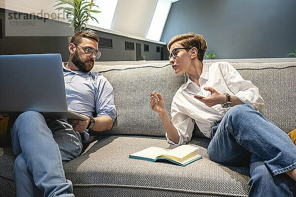 Männliche und weibliche Geschäftskollegen diskutieren  während sie auf einem beleuchteten Sofa in Coworking-Büroräumen sitzen