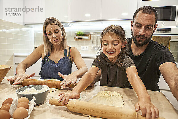 Eltern mit lächelnder Tochter kneten Pizzateig auf dem Tisch in der Küche