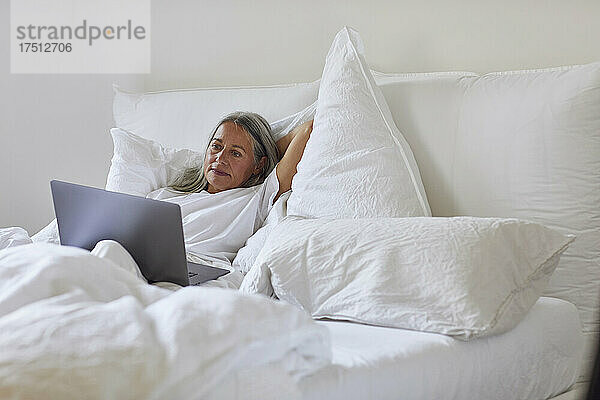 Reife Frau benutzt Laptop  während sie zu Hause im Bett liegt