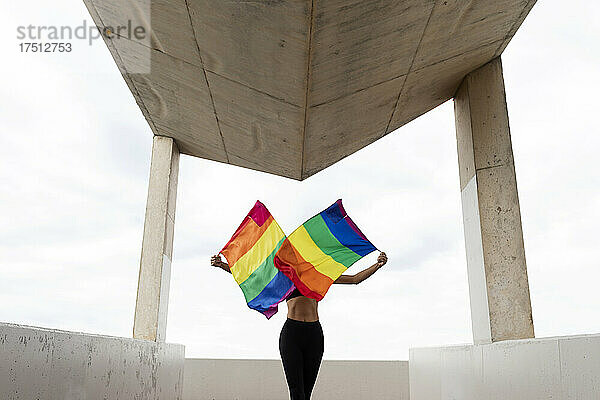 Eine Frau mittleren Alters schwenkt Regenbogenfahnen  während sie auf einem Bauwerk vor dem Himmel steht