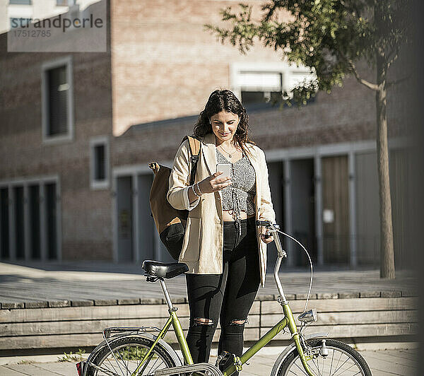 Junge Frau benutzt Smartphone  während sie mit dem Fahrrad auf der Stadtstraße steht