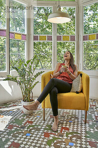 Fröhliche Frau mit Tasse sitzt zu Hause auf einem Sessel am Fenster