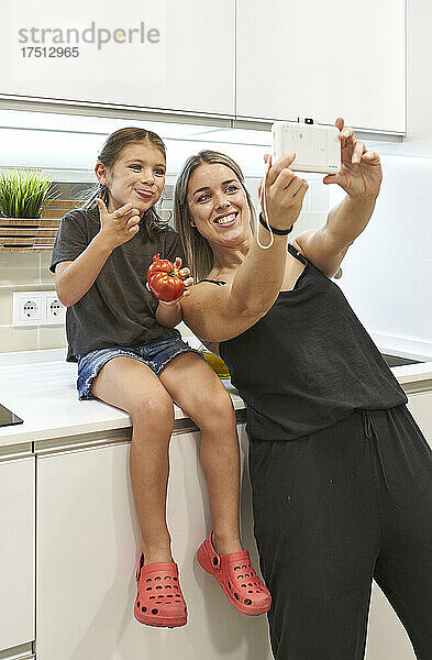 Glückliche Mutter macht ein Selfie mit ihrer Tochter  die zu Hause auf der Küchentheke Tomaten isst