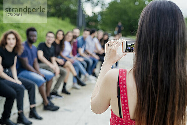 Frau fotografiert Freunde  die mit Smartphone im Park in einer Reihe sitzen