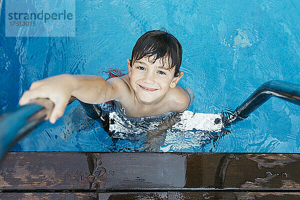 Lächelnder Junge hält Leiter  während er im Schwimmbad steht