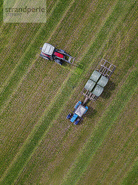 Luftaufnahme von zwei Traktoren  die Heuballen auf dem Feld sammeln