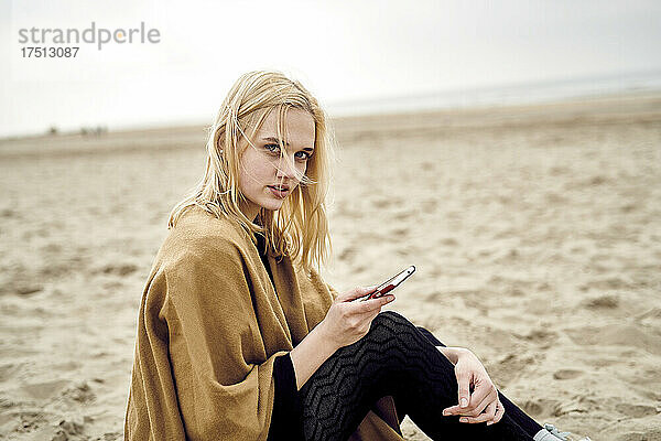 Niederlande  Porträt einer blonden jungen Frau mit Smartphone am Strand sitzend