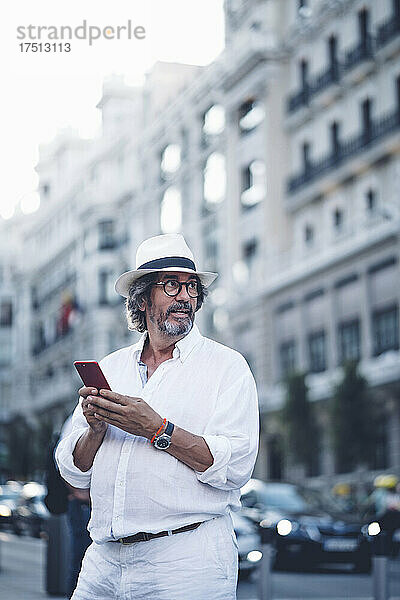 Attraktiver älterer Mann mit weißer Kleidung und Hut schaut weg  während er sein Smartphone in der Straße von Madrid hält