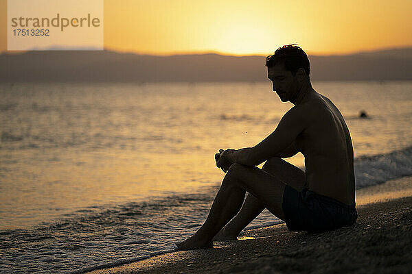 Nachdenklicher Mann mittleren Alters mit nacktem Oberkörper  der bei Sonnenuntergang am Ufer vor dem Himmel sitzt