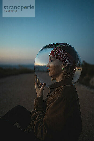 Woman with a fish bowl on her head sitting on a road in the countryside at dusk