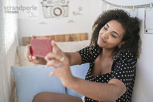 Junge Frau mit lockigem Haar macht ein Selfie mit dem Smartphone  während sie zu Hause an der Wand sitzt
