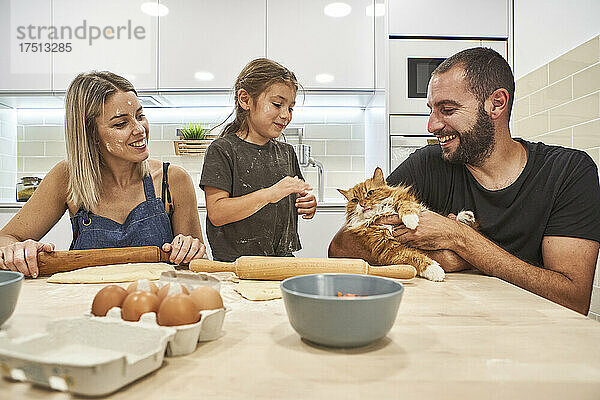 Mutter knetet Teig mit Nudelholz  während Vater und Tochter mit Katze auf dem Küchentisch spielen