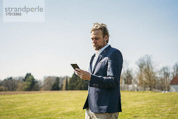 Geschäftsmann benutzt Smartphone  während er an einem sonnigen Tag im Park vor klarem Himmel steht