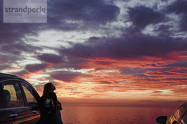 Frau mittleren Alters steht bei Sonnenuntergang mit dem Auto am Strand vor bewölktem Himmel