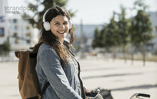 Lächelnde Studentin  die an einem sonnigen Tag in der Stadt steht und über Kopfhörer Musik hört