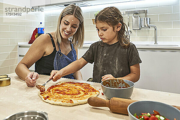 Mutter schaut Tochter dabei zu  wie sie in der Küche Soße auf Pizzateig aufträgt