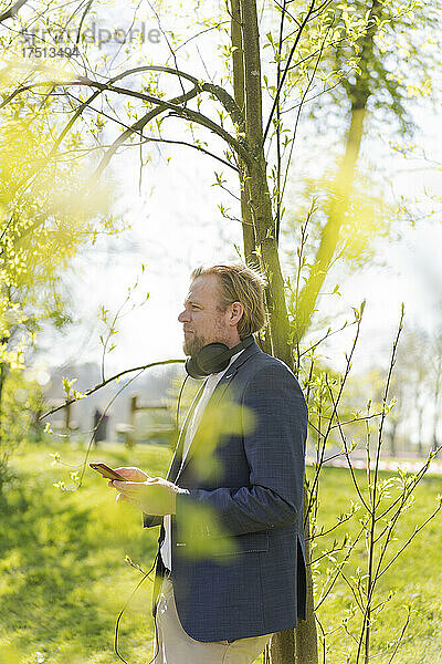 Nachdenklicher männlicher Berufstätiger schaut weg  während er an einem sonnigen Tag mit dem Smartphone gegen einen Baum im Park steht